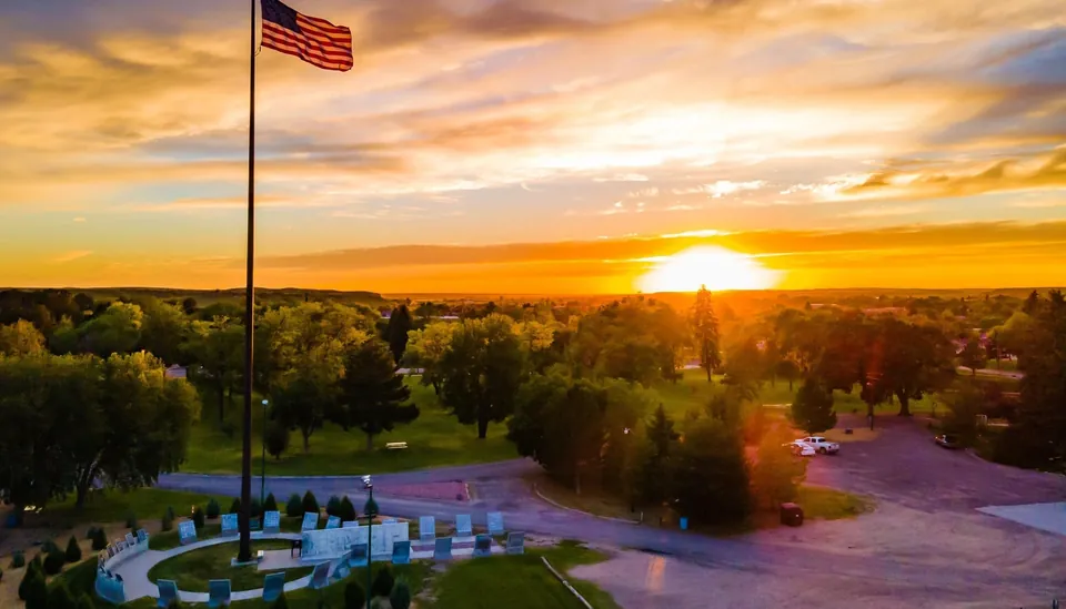 An American flag waves over a monument in a park against a setting sun and over the treetops in the distance.