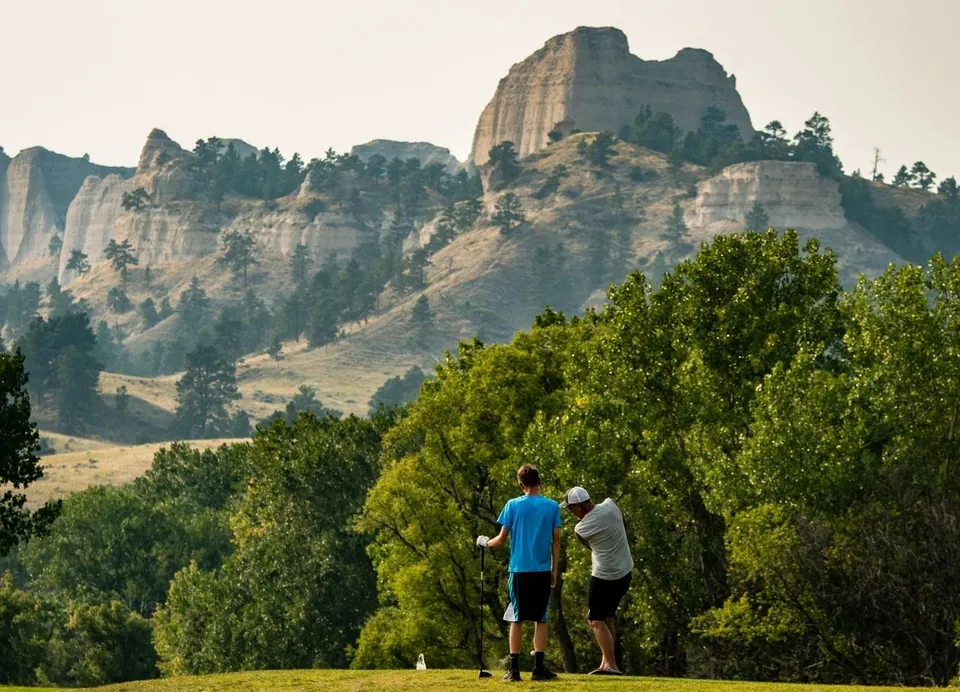 Two men golf and a towering rock formation rises up above them in the background.